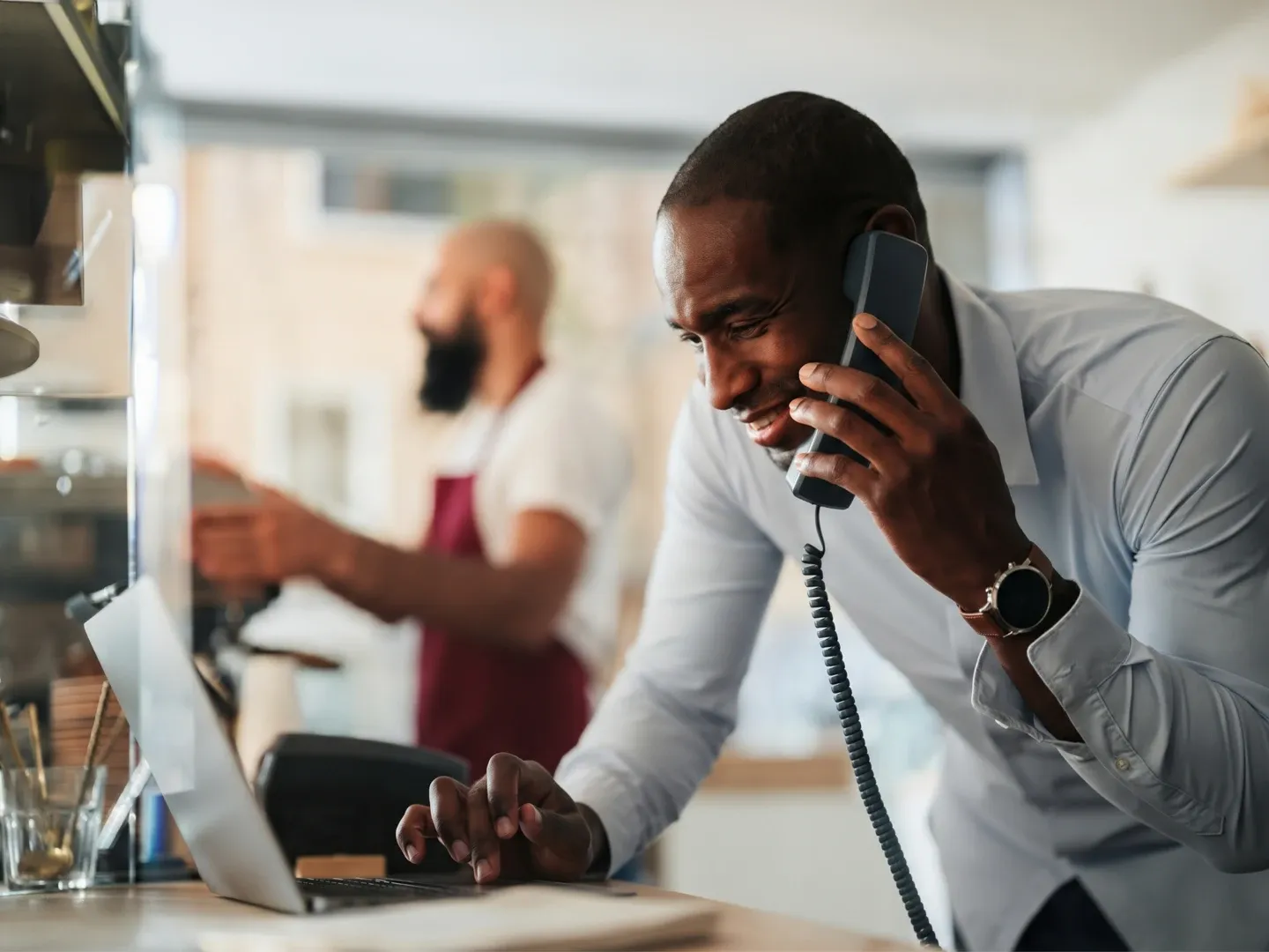 Small business owner speaking on a landline phone while working on a laptop at a cafe counter.